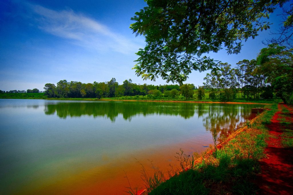 Lago de águas calmas refletindo árvores e céu azul, com margem de terra vermelha, no Parque Maria Tuca, em Tatuí