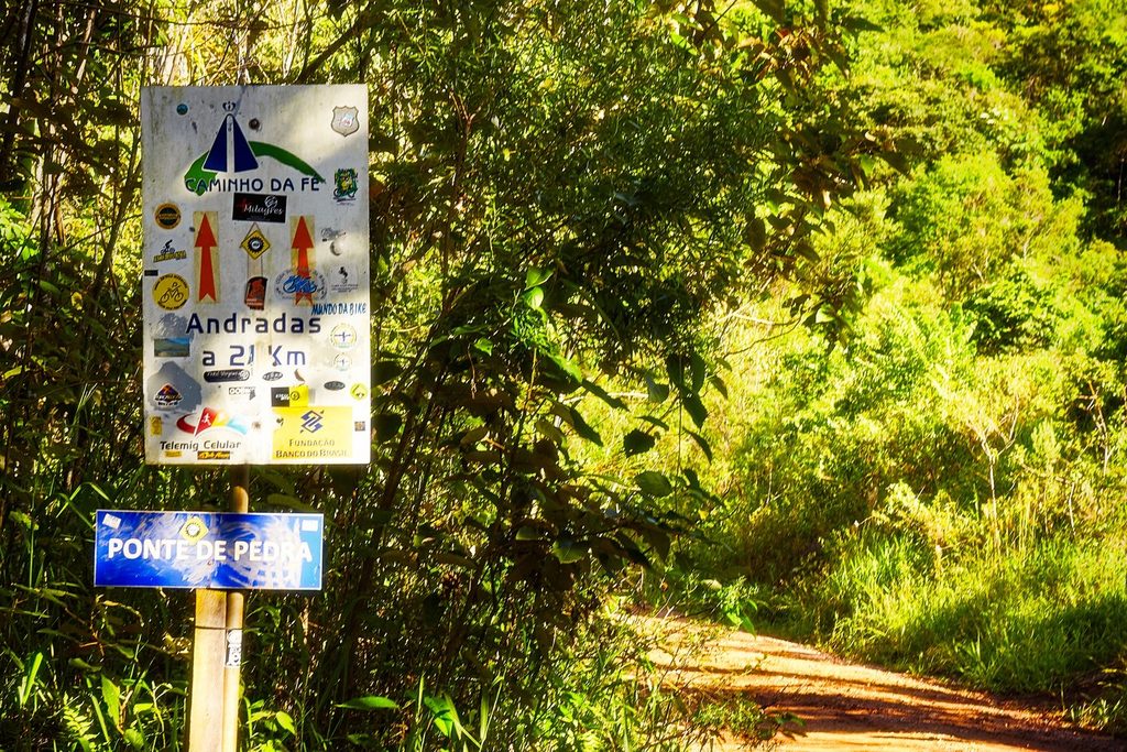 Placa do Caminho da Fé em Águas da Prata indicando Andradas a 21 km, ao lado de uma estrada de terra no meio da mata