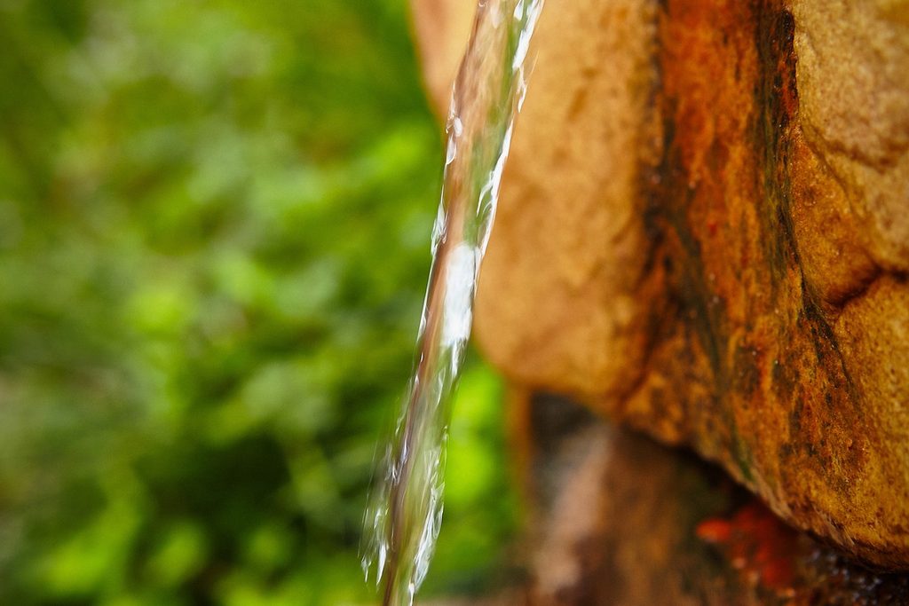 Fio de água mineral caindo de uma bica de pedra avermelhada, com mata verde desfocada ao fundo