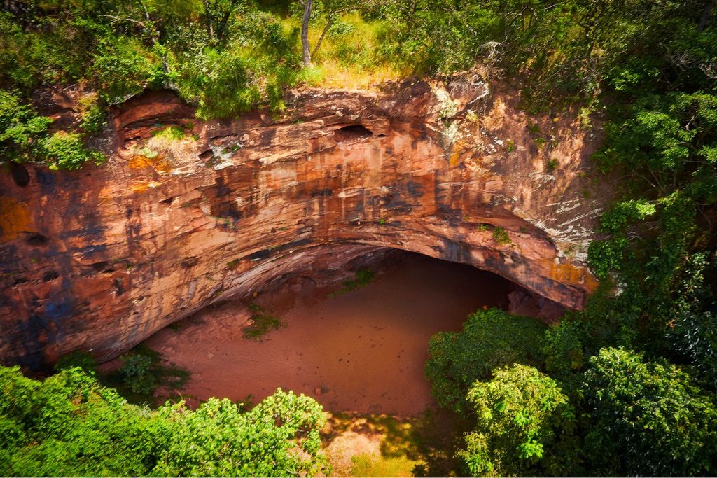 Grande boca da Gruta do Itambé vista de cima, com paredão de pedra avermelhada cercado de mata