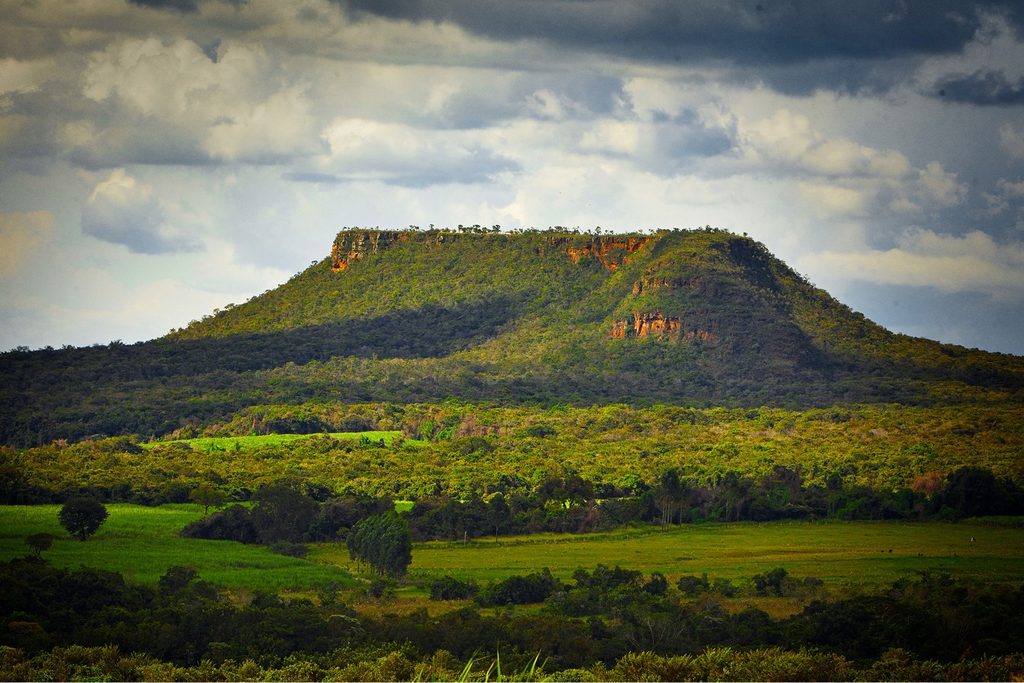 Morro de topo achatado coberto de vegetação, com paredões de pedra e campos verdes em volta