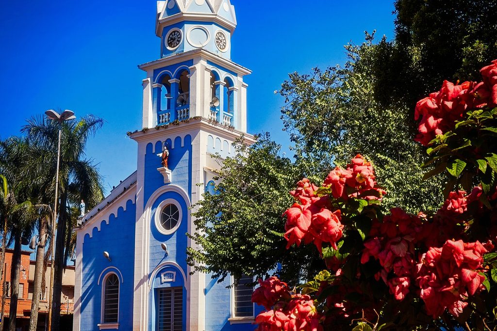Torre azul e branca da Igreja Matriz com relógio, palmeiras e flores vermelhas em primeiro plano