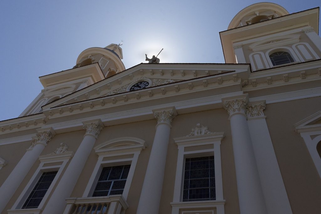 Fachada clara da Catedral de Nossa Senhora do Amparo vista de baixo, com colunas, frontão com relógio e duas torres contra o céu azul