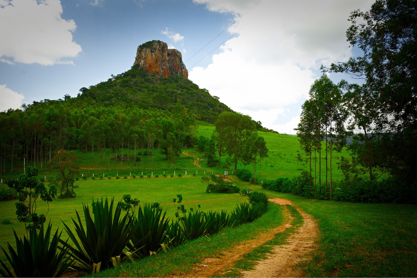 Morro de arenito com topo rochoso sobre encosta de pasto verde e eucaliptos, com estrada de terra em primeiro plano, em Analândia
