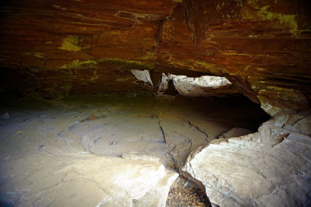 Interior de gruta de arenito com teto em camadas avermelhadas e chão de rocha clara iluminado pela luz de uma abertura