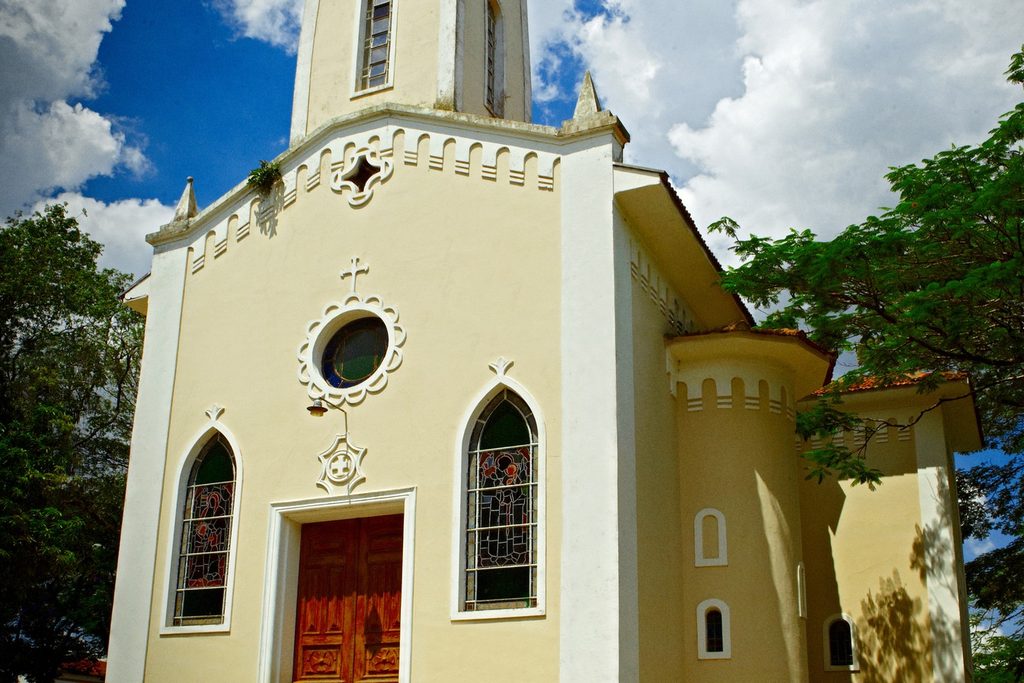 Fachada creme da Igreja Matriz de Analândia com torre, janelas em arco e porta de madeira vermelha