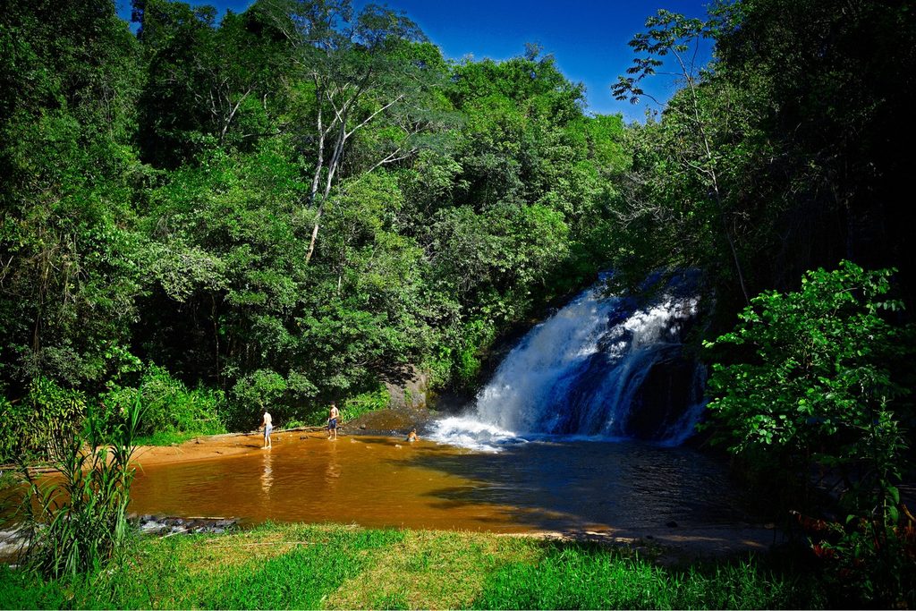 Cachoeira branca descendo entre a mata para um poço de água avermelhada, com banhistas na margem, no Salto Major Levy, em Analândia