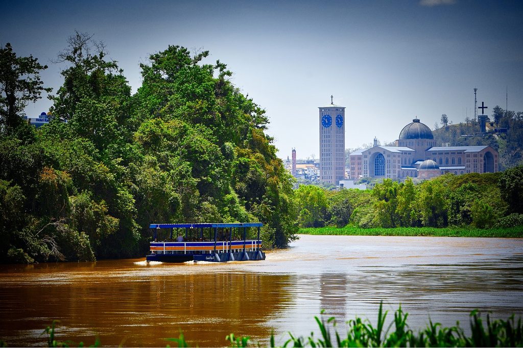 Balsa de passeio navegando no rio Paraíba do Sul, com mata na margem e o Santuário Nacional de Aparecida ao longe
