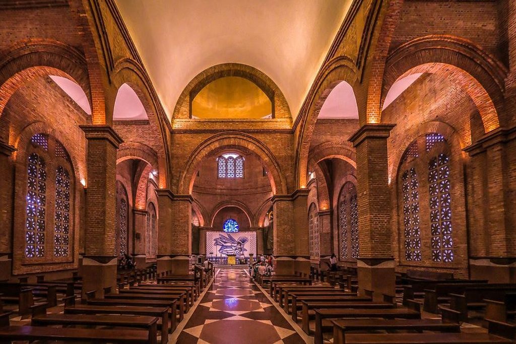 Nave com bancos de madeira, pilares e arcos de tijolos aparentes e vitrais azuis, com o altar ao fundo