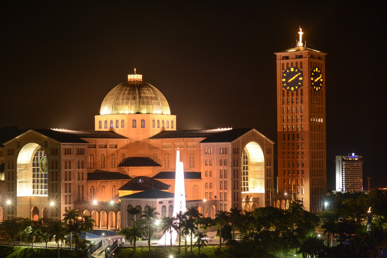 Santuário Nacional de Aparecida iluminado à noite, com a cúpula dourada, a torre do relógio e uma fonte iluminada à frente