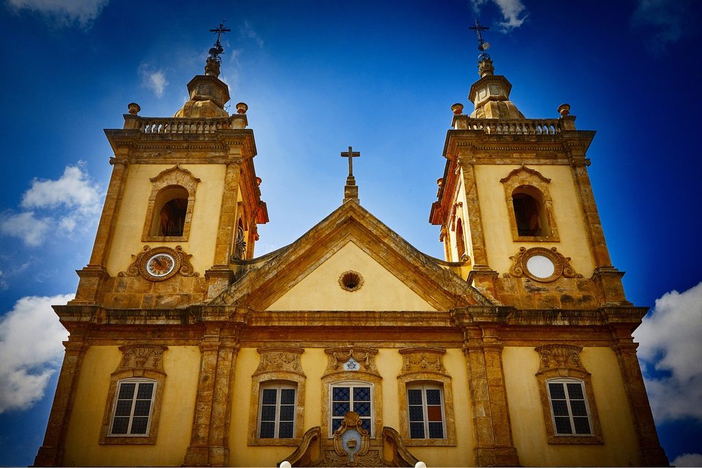Fachada amarela e branca da Basílica Velha de Aparecida, com duas torres com relógio e cruzes, sob céu azul