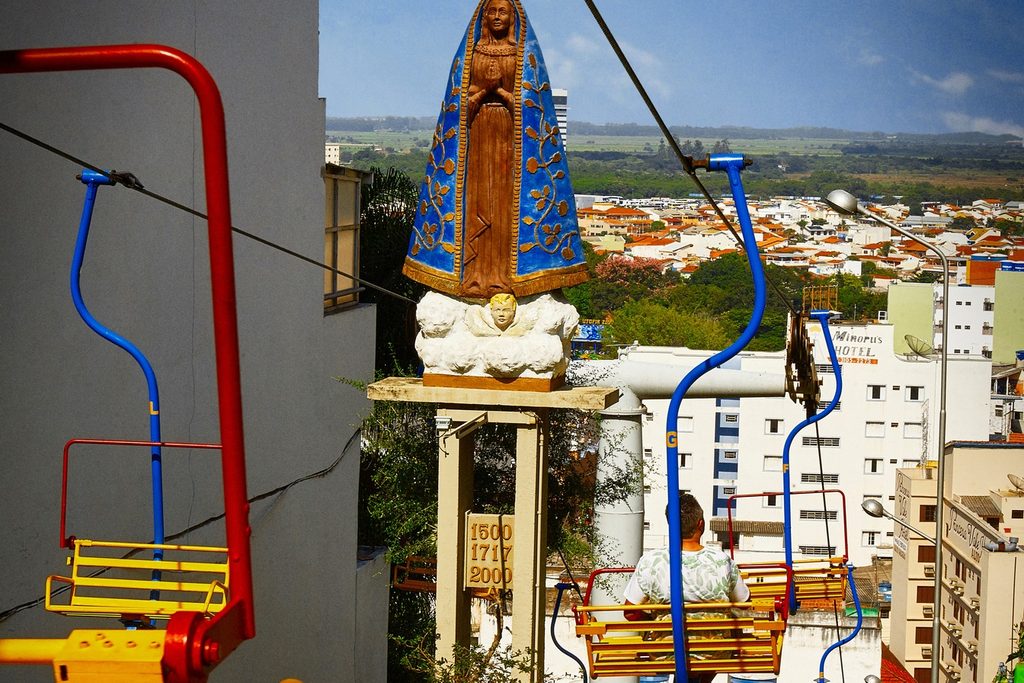Cadeirinhas coloridas de teleférico passando ao lado de uma imagem de Nossa Senhora, com os telhados da cidade ao fundo