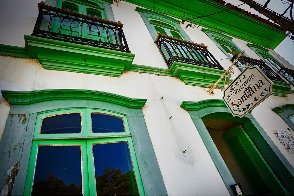 Fachada branca do Hotel Sant'Ana, em Areias, com janelas e sacadas de ferro emolduradas em verde e a placa do hotel e restaurante