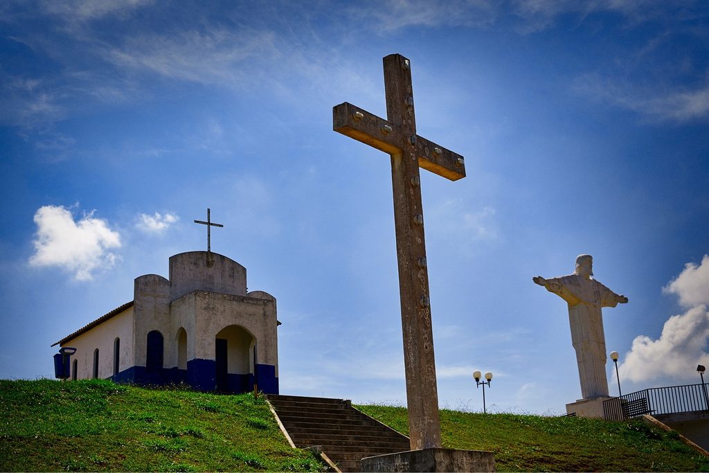 Capelinha branca, cruzeiro de madeira e estátua do Cristo no alto do morro do mirante, sob céu azul com nuvens