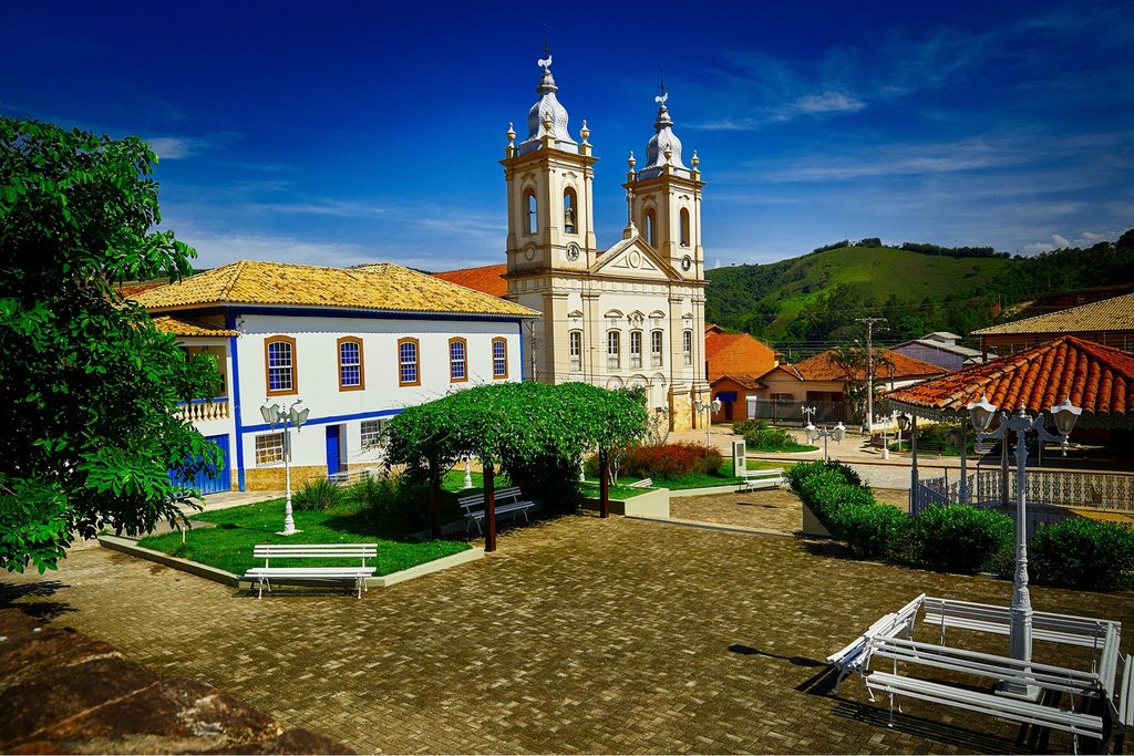 Praça Nove de Julho com bancos e jardim, casarão branco e azul e as duas torres da Igreja Matriz de Sant'Ana ao fundo