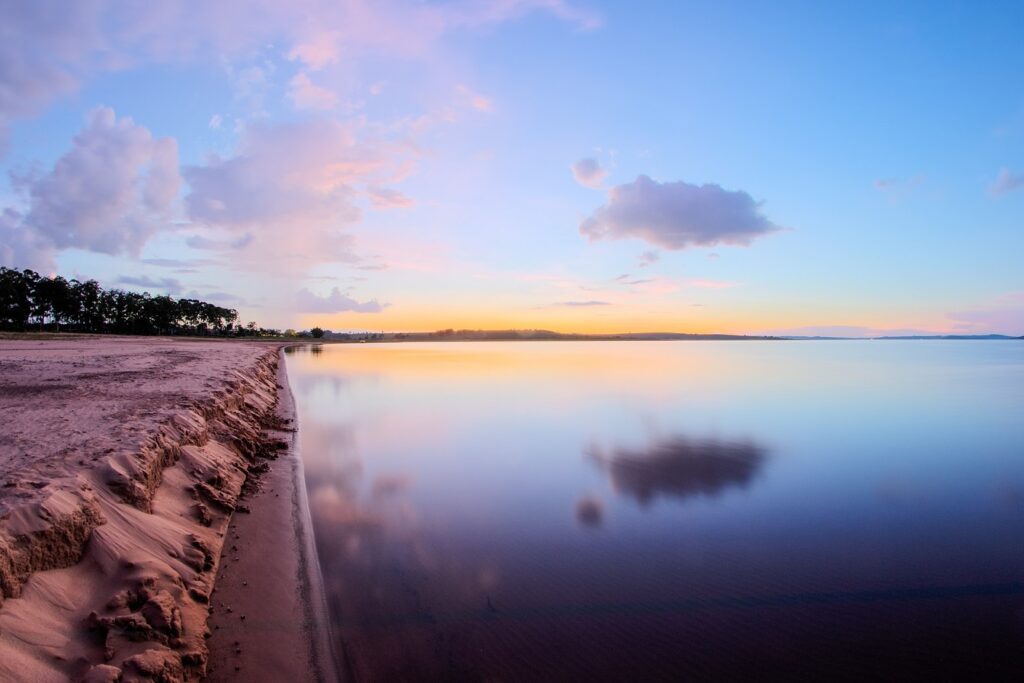 Margem de areia da represa em Avaré ao entardecer, com a água parada refletindo nuvens cor-de-rosa e a mata no horizonte