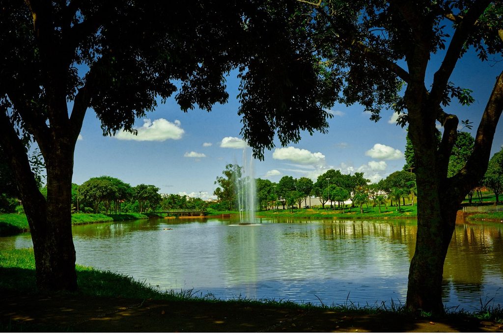 Lago com chafariz cercado de árvores e gramado sob céu azul em Batatais