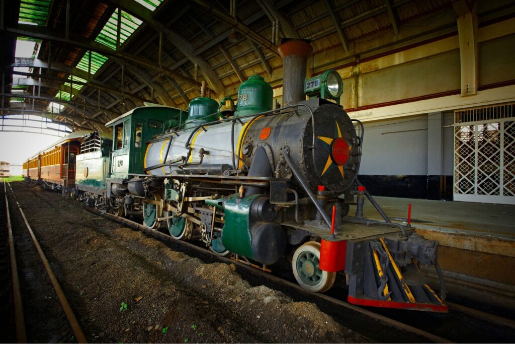 Locomotiva a vapor preta e verde com estrela vermelha na frente, parada nos trilhos sob a cobertura de uma antiga estação, no Museu Ferroviário de Bauru