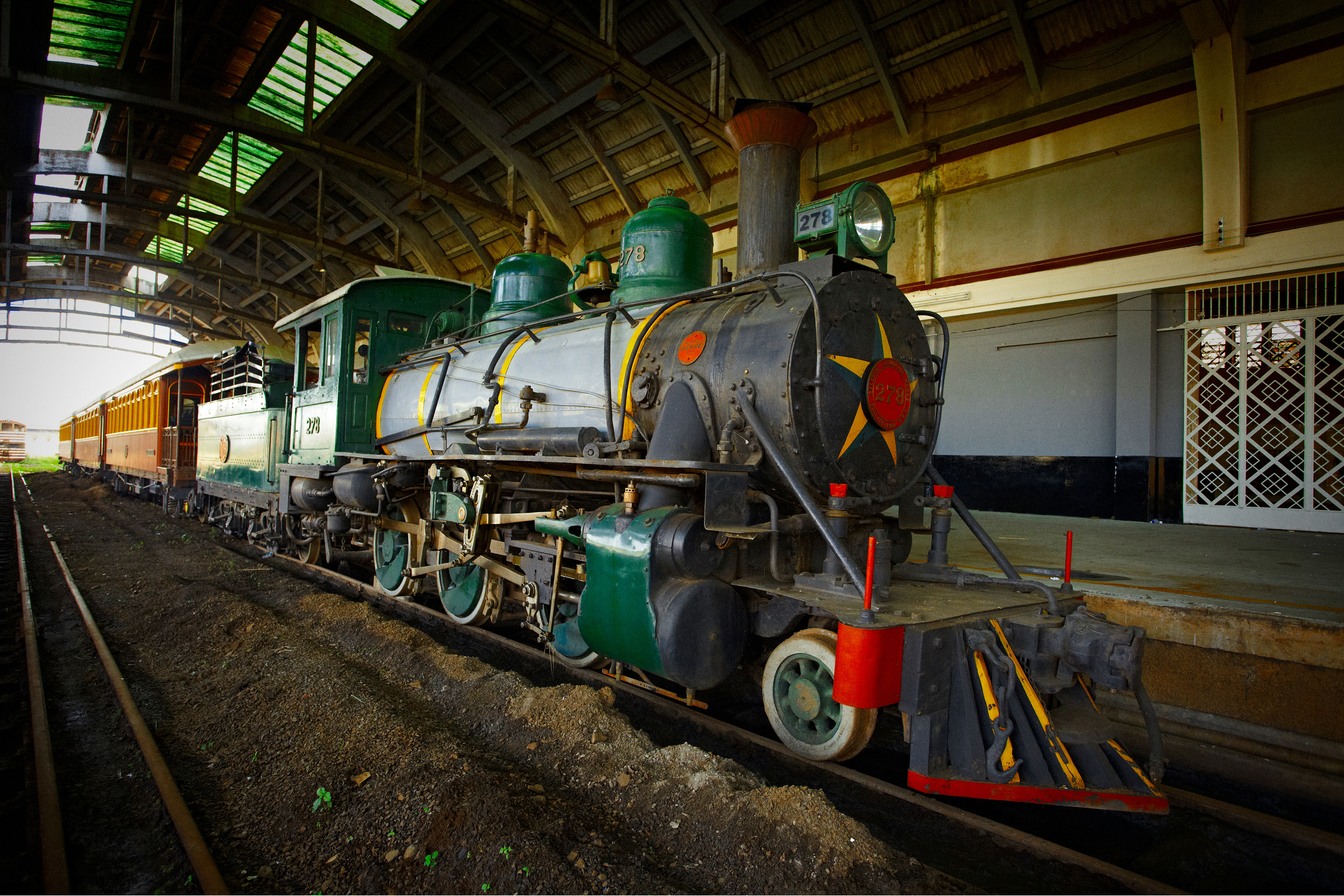 Locomotiva a vapor preta e verde com estrela vermelha na frente, parada nos trilhos sob a cobertura de uma antiga estação, no Museu Ferroviário de Bauru