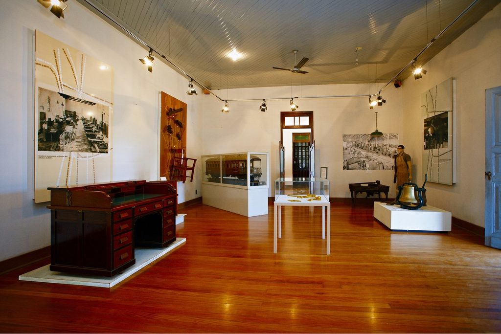 Sala de exposição com piso de madeira, móveis antigos, vitrines e fotografias da ferrovia nas paredes, no Museu Ferroviário de Bauru