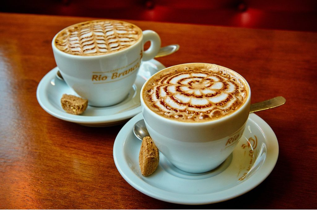 Duas xícaras de cappuccino com desenho na espuma sobre uma mesa de madeira na Cafeteria Rio Branco