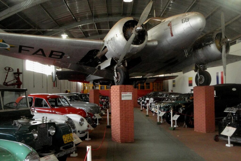 Avião bimotor prateado da FAB suspenso sobre pilares de tijolo, acima de fileiras de carros antigos, no salão do museu de Bebedouro