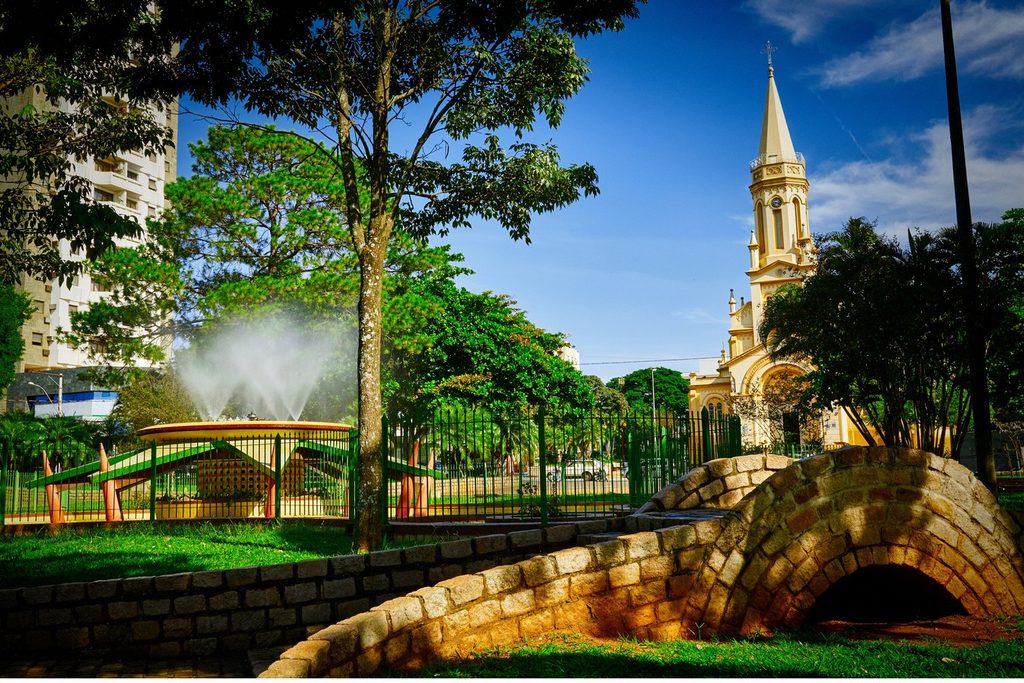 Praça arborizada com chafariz, ponte de pedra e a torre alta da igreja matriz ao fundo, sob céu azul