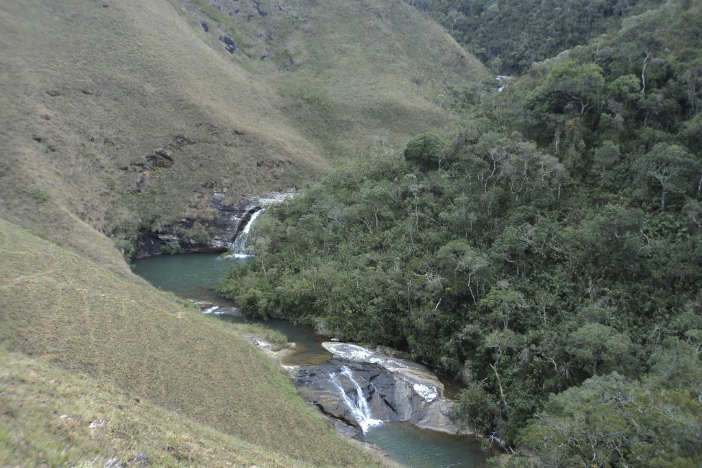 Rio formando pequenas quedas e um poço verde num vale entre campos de altitude e mata