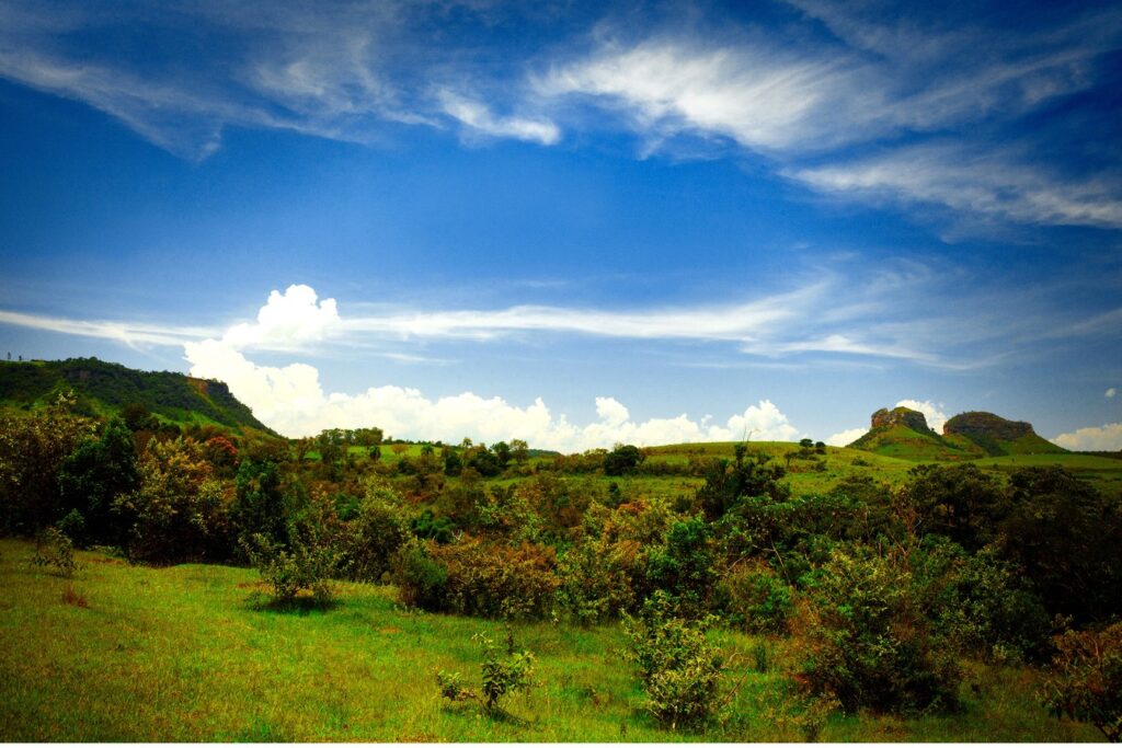 Vale do Gigante, em Bofete, com pasto e mata em primeiro plano e as Três Pedras no horizonte sob céu azul com nuvens