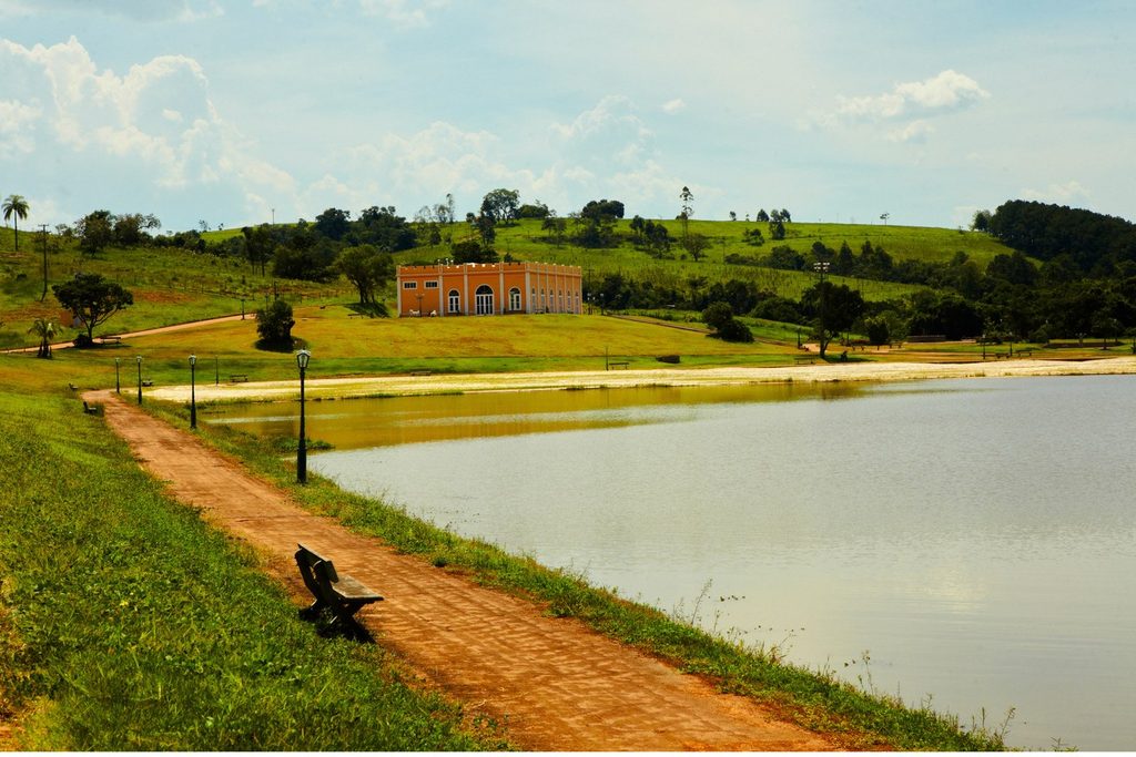 Lago com caminho de tijolos e faixa de areia no Espaço Castelo, com um casarão ao fundo