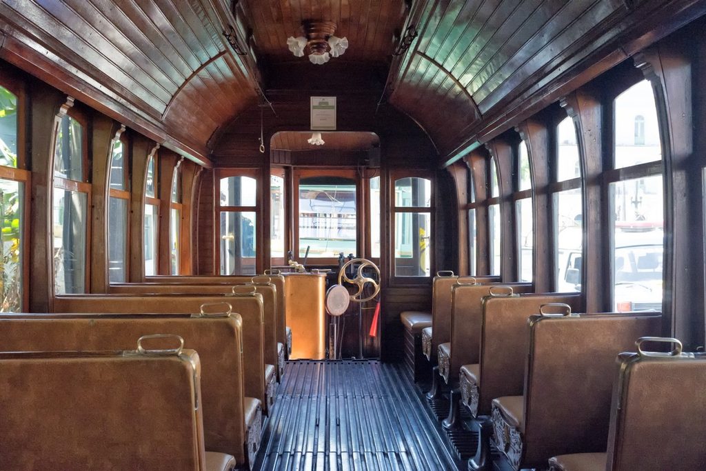 Interior de um bonde antigo com teto e paredes de madeira, bancos de couro e o comando do motorneiro ao fundo