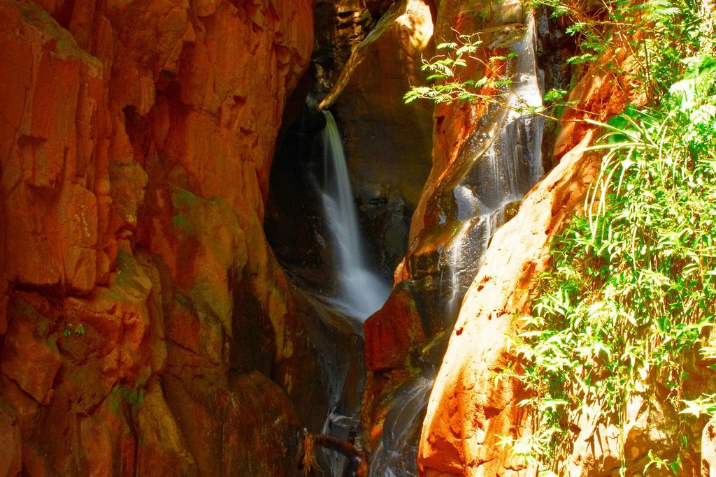 Cachoeira fina caindo entre paredões de pedra avermelhada e vegetação, no Vale da Indiana