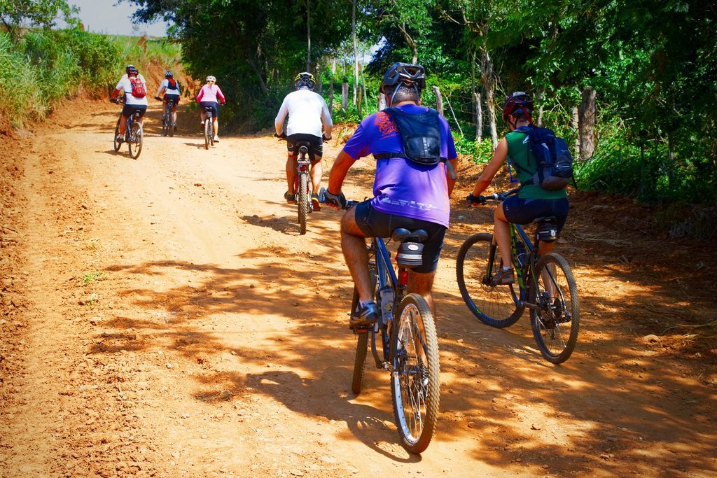 Grupo de ciclistas pedalando numa estrada de terra vermelha entre a mata, na zona rural de Botucatu