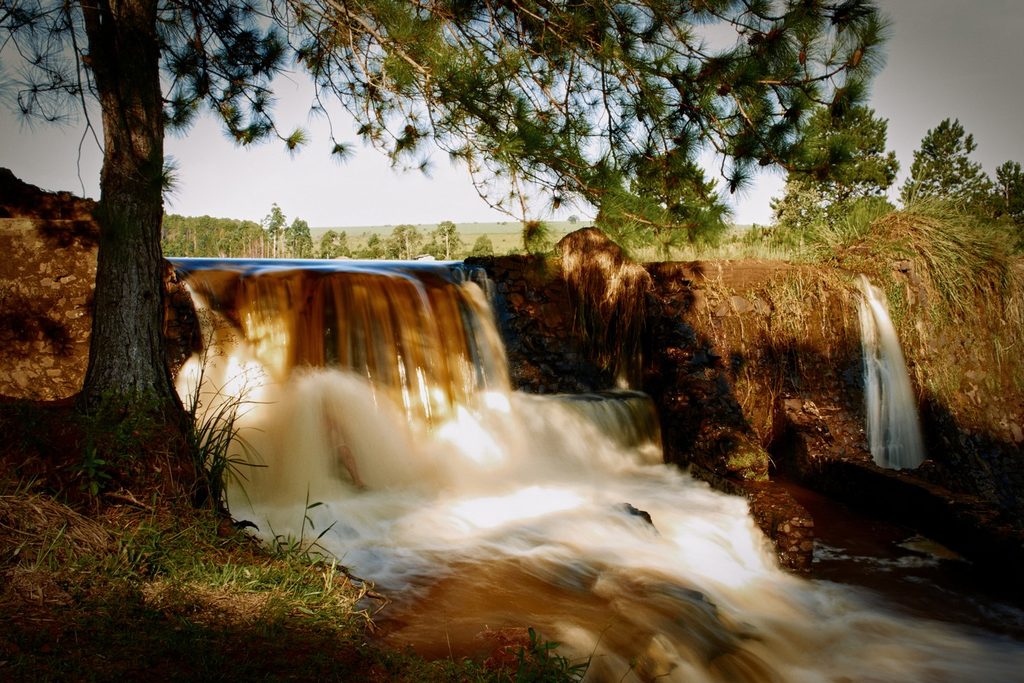 Cachoeira Véu de Noiva, em Botucatu, com água cor de chá descendo em degraus largos entre pedras e um pinheiro