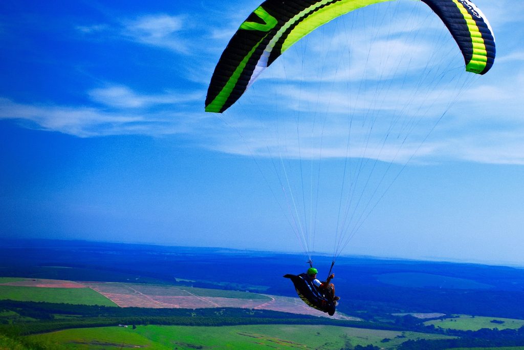 Parapente verde e preto voando alto sobre o vale e as plantações da Cuesta, sob céu azul