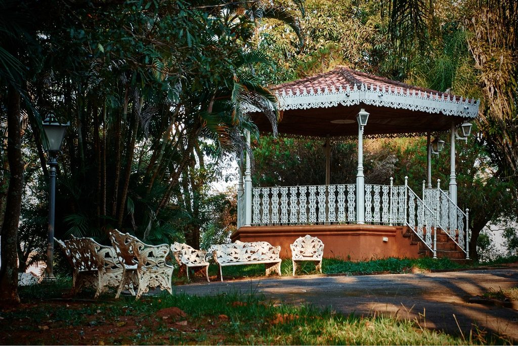Coreto branco rendado com cadeiras de ferro em volta, entre árvores do Jardim Público de Bragança Paulista