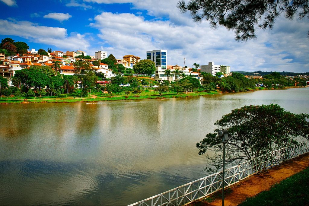Lago do Taboão com a água refletindo o céu, gradil branco na margem e prédios e casas da cidade ao fundo