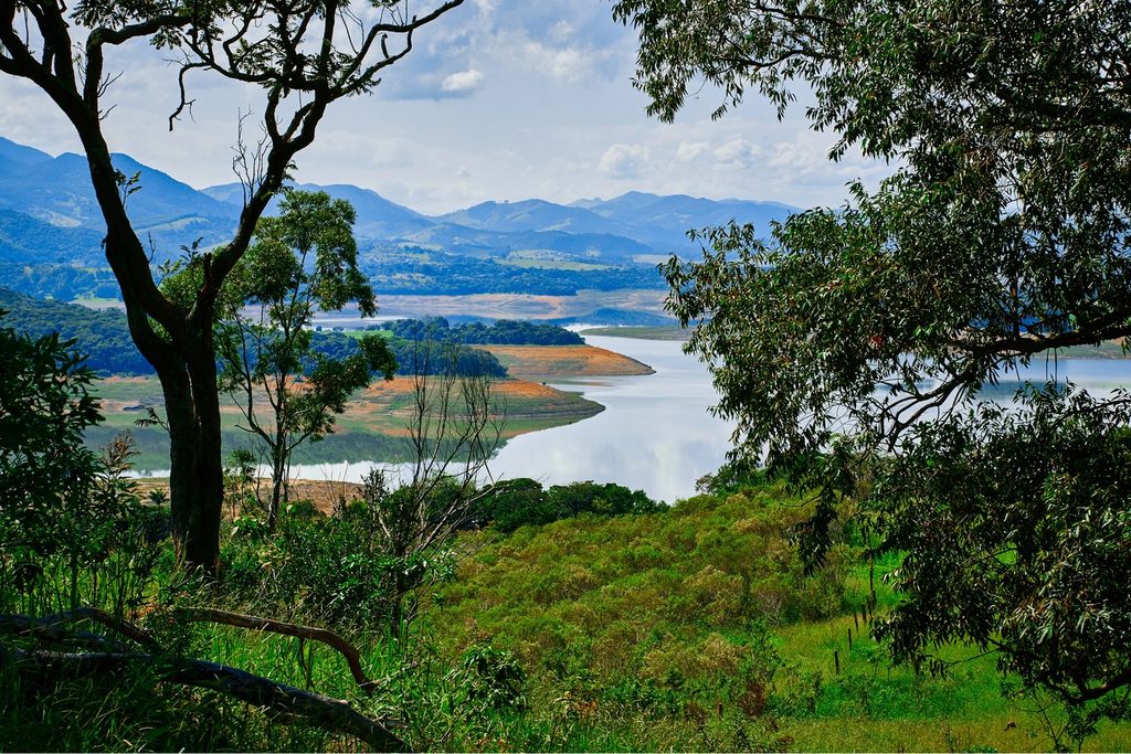 Represa Jaguari-Jacareí entre morros verdes e montanhas azuladas, vista do alto por entre árvores, em Bragança Paulista