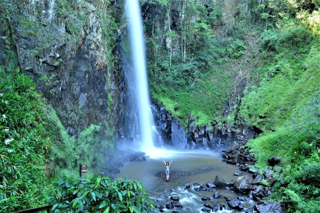 Cachoeira alta e fina caindo de um paredão coberto de mata num poço raso, com uma pessoa de braços abertos lá embaixo