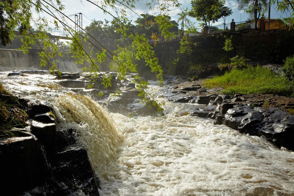 Queda d'água e corredeiras do rio Jacaré-Pepira entre pedras escuras, com ponte pênsil e árvores ao fundo, no Parque dos Saltos