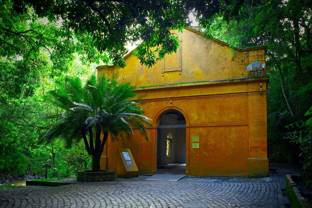 Prédio histórico amarelo com porta em arco, cercado de mata e palmeira, no Parque dos Saltos, em Brotas