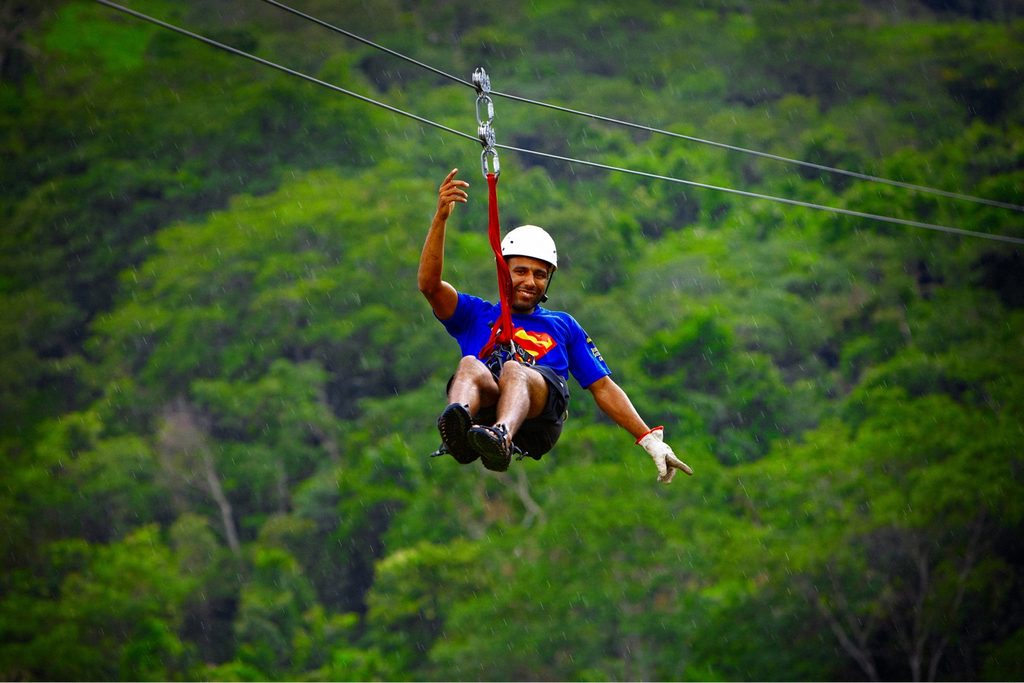 Homem de capacete atravessando de tirolesa por cima da mata, de braços abertos, em Brotas