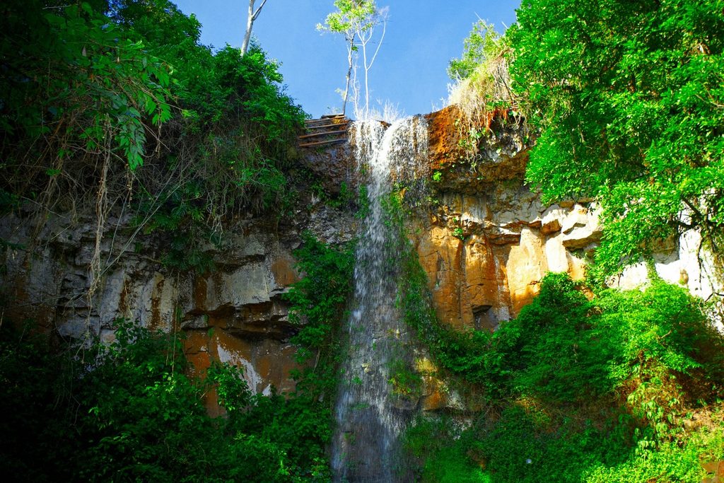 Cachoeira fina descendo por um paredão de pedra alaranjada no meio da mata, no Sítio Três Quedas