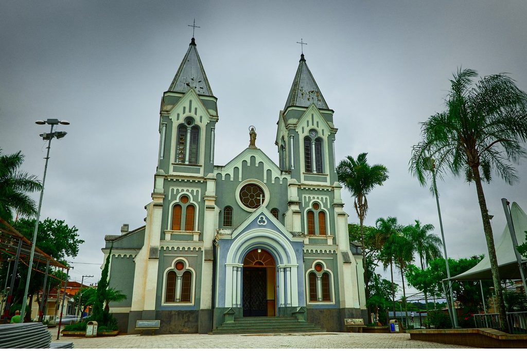 Basílica com duas torres pontiagudas e fachada cinza e branca, ao lado de palmeiras