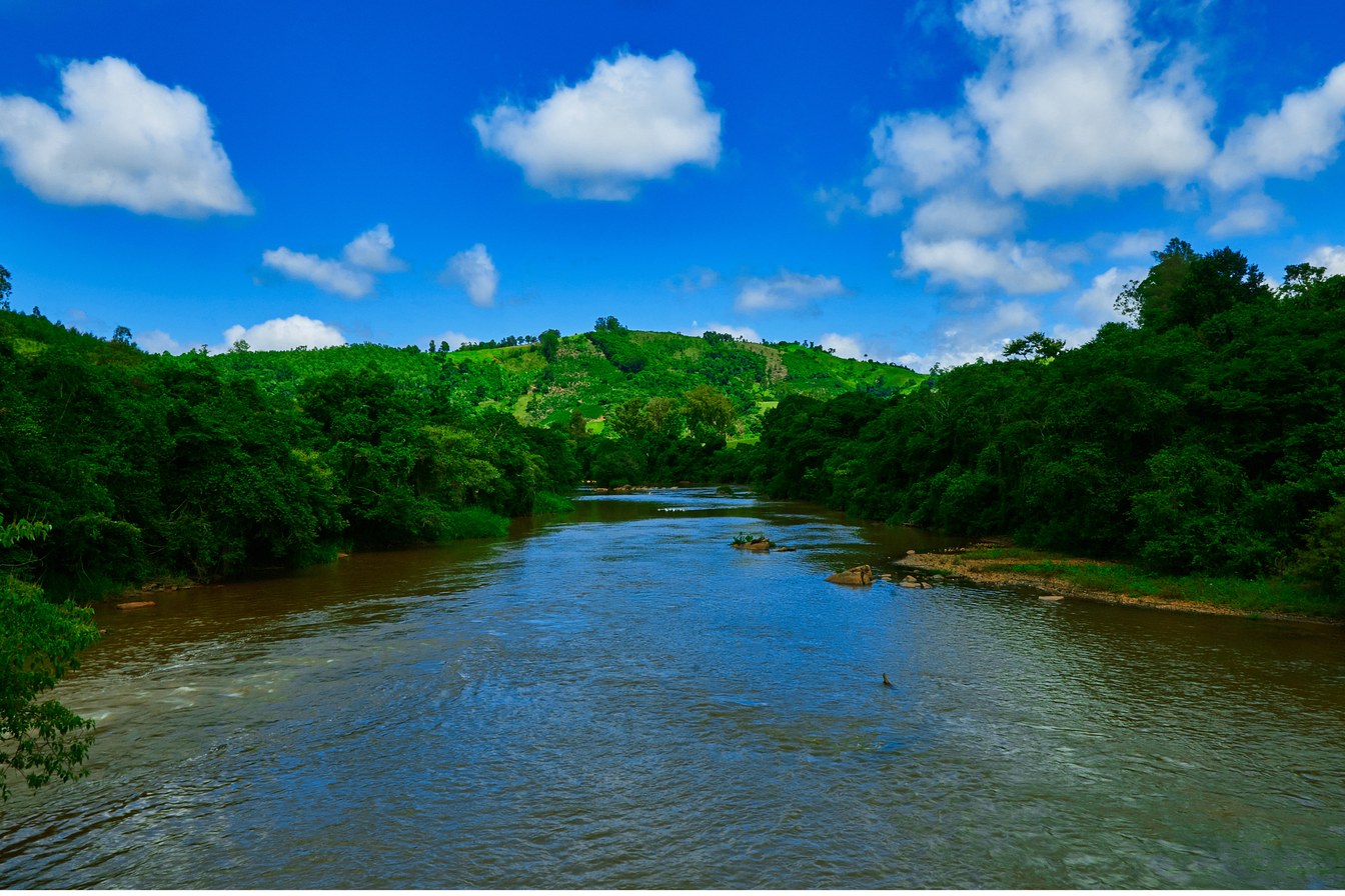 Rio Pardo em Caconde correndo entre margens de mata e morros verdes, sob céu azul com nuvens