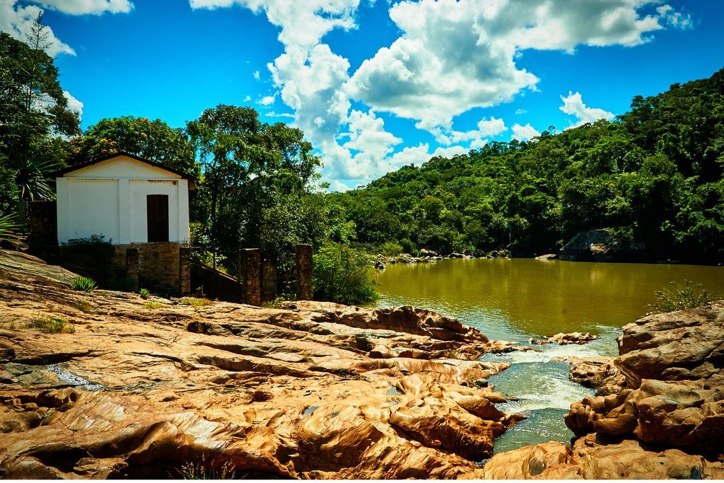 Pequeno prédio branco da antiga usina sobre as rochas, com o rio e a mata ao fundo