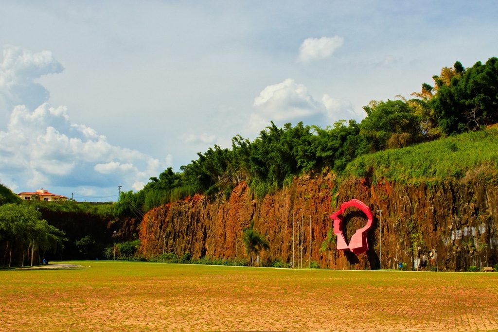 Paredão de pedra avermelhada da antiga pedreira, com mata no alto, gramado e uma escultura vermelha, na Pedreira do Chapadão