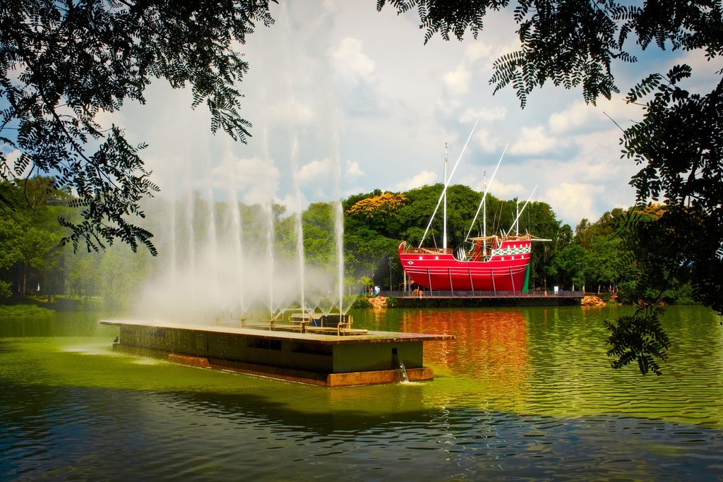 Lagoa do Taquaral, em Campinas, com chafariz de jatos de água e a réplica de caravela vermelha atracada num deck entre árvores