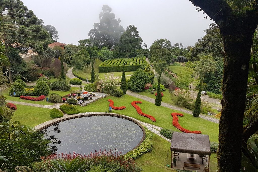 Jardins com espelho d'água redondo, canteiros de flores vermelhas em curva, labirinto de arbustos e morros com neblina ao fundo, no Parque Amantikir, em Campos do Jordão