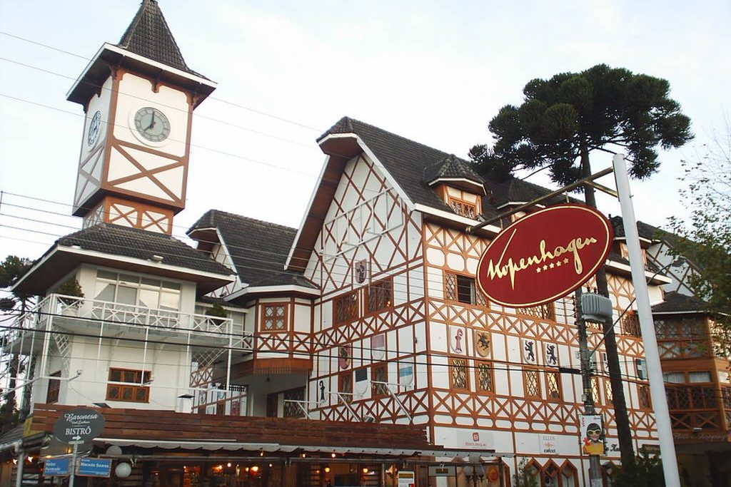 Casarões em estilo enxaimel brancos e marrons com torre de relógio e uma araucária ao fundo, no centro comercial do Capivari